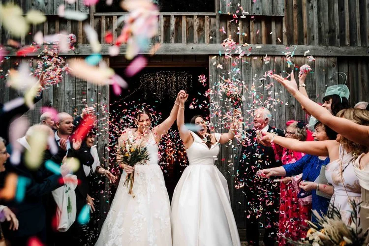 A joyful confetti aisle moment captured by Amy Faith Photography as two brides celebrate hand-in-hand outside a rustic barn venue. Colourful confetti fills the air while guests cheer and throw handfuls into the scene. One bride carries a wild bouquet