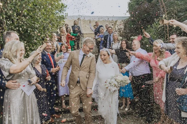 A joyful and celebratory confetti moment captured by YU Photography, showing the newly married couple walking hand in hand through a pathway of cheering guests. Colourful petals fill the air as friends and family shower them with excitement, creating