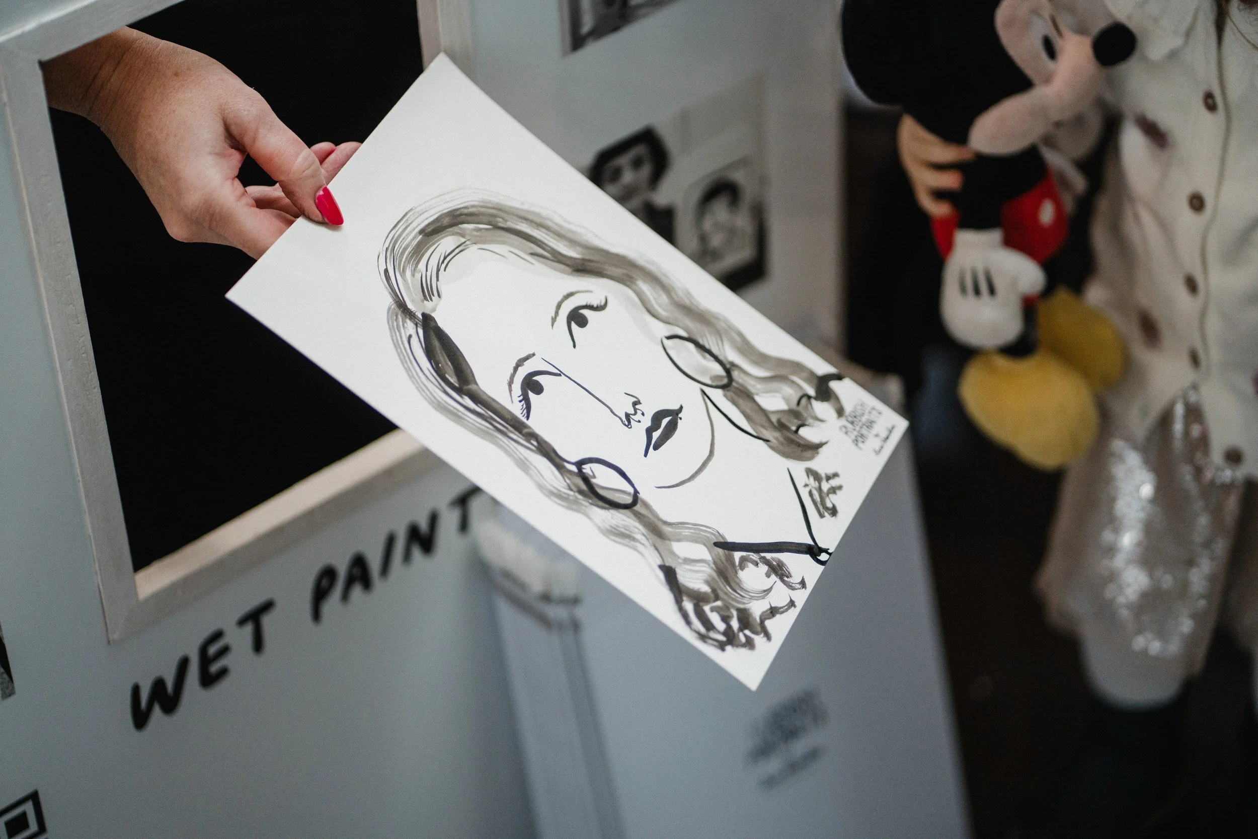 Close-up of a freshly created black and white portrait being handed out from the Rubbish Portraits live illustration booth. The drawing, featuring a stylish woman with wavy hair and hoop earrings, is presented by an artist with bright red nails, whil