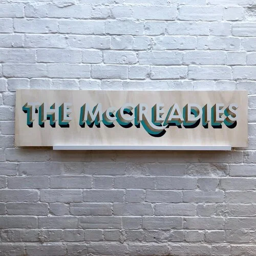 A bespoke wooden sign by Esther of Sylvie Signs featuring the hand-painted phrase “THE McCREADIES” in bold 3D teal and cream lettering. The design has a soft natural wood base with depth and shadow detail that gives the typography a vintage yet moder