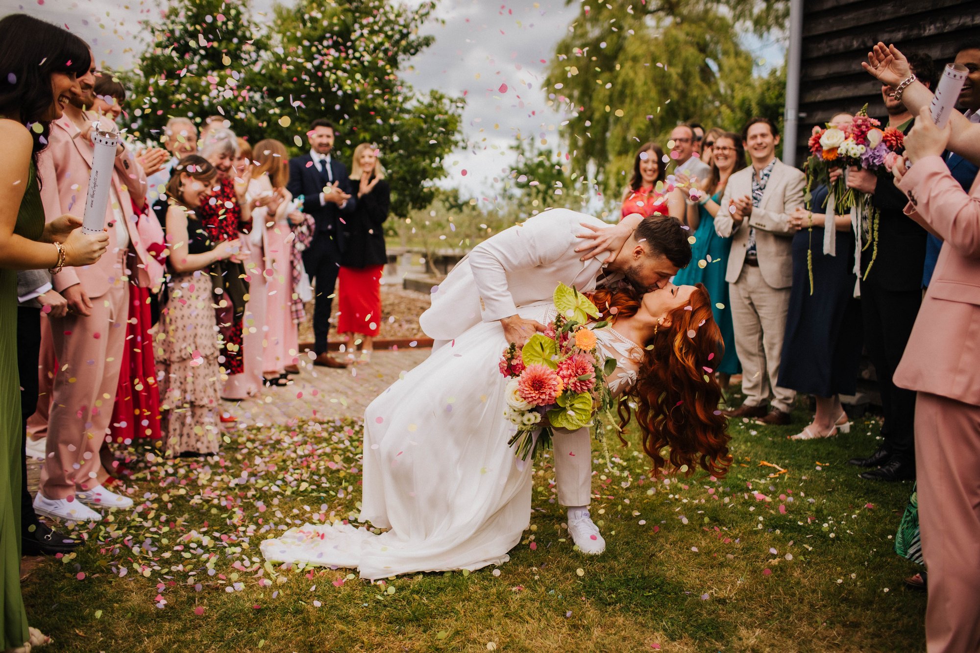 A colourful shot of a modern bride and groom kissing outside The Canary Shed. Their guests are covering them in confetti.