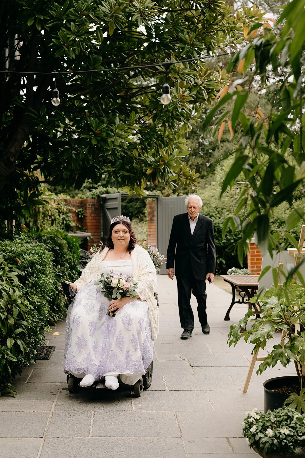 A disabled bride in a beautiful, lilac wedding dress is about to make her way down the aisle with her father who is wearing a a black suit. She's holding a lilac bridal bouquet and her wheelchair has matching florals attached to the back of it.