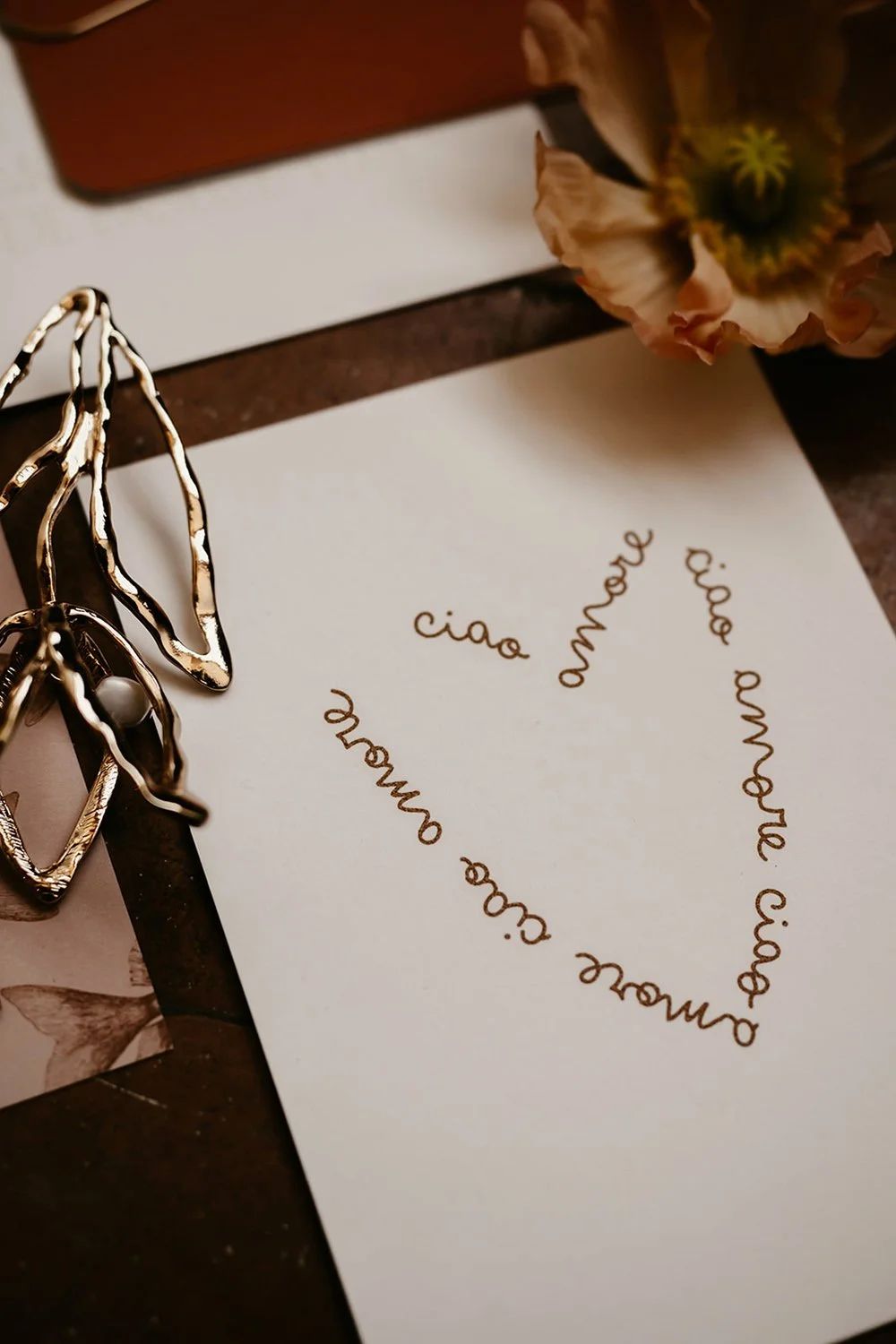 A refined wedding stationery detail featuring an ivory card printed with the romantic Italian phrase ciao amore arranged in a looping heart shape styled alongside organic gold jewellery and a soft peach flower creating a warm modern editorial look pe