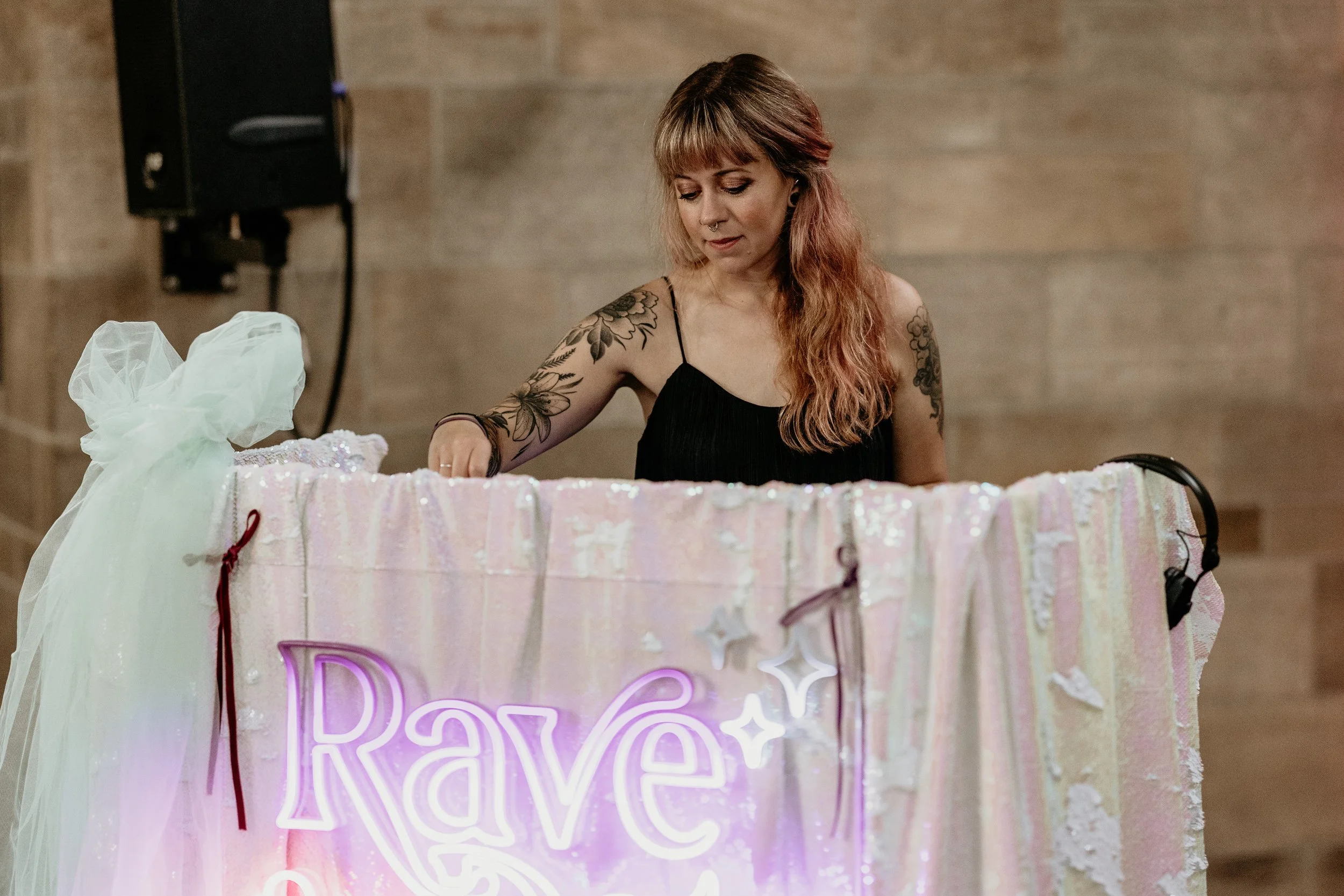 This photo shows Rave The Date DJ Cara in action, focused behind her shimmering pastel and sequin DJ booth adorned with the glowing “Rave the Date” sign. With her headphones nearby and soft lighting highlighting the scene, Cara’s calm confidence and 