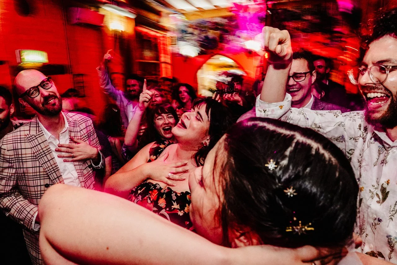 A crowd of guests pack a vibrant, neon lit dance floor with arms raised, faces flushed from dancing and laughter, one woman at the centre clutching her chest in joyful disbelief while friends cheer around her and motion blurs streak the background wi