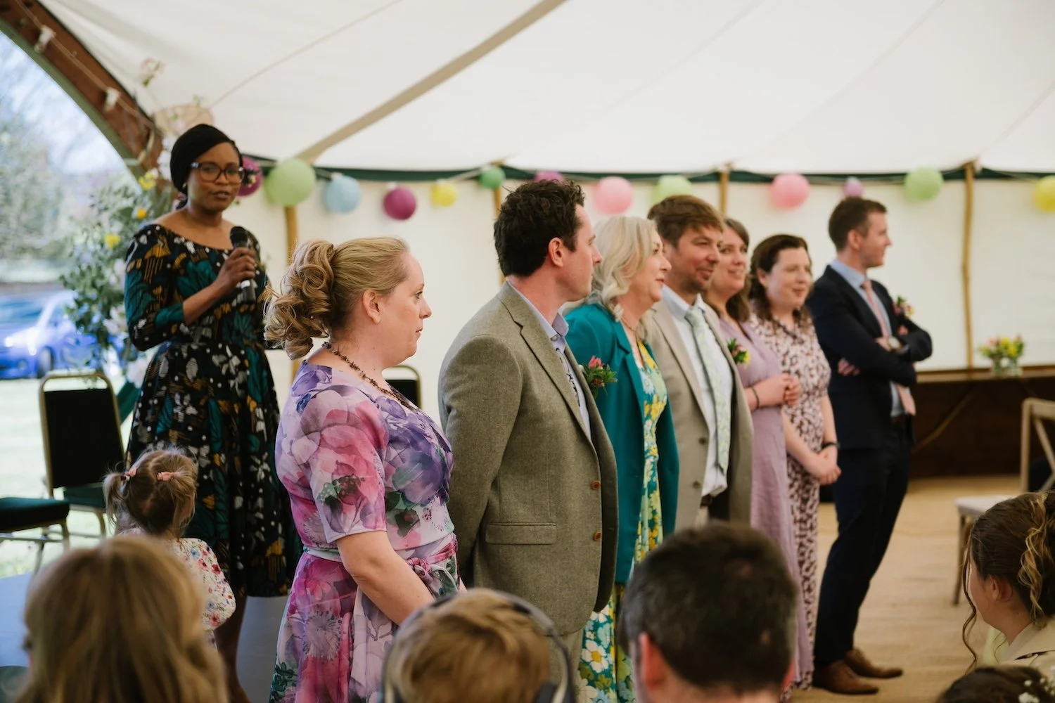 A wedding ceremony with guests all in a row. There is a Black lady in the background holding a microphone. She is wearing glasses and a floral dress.