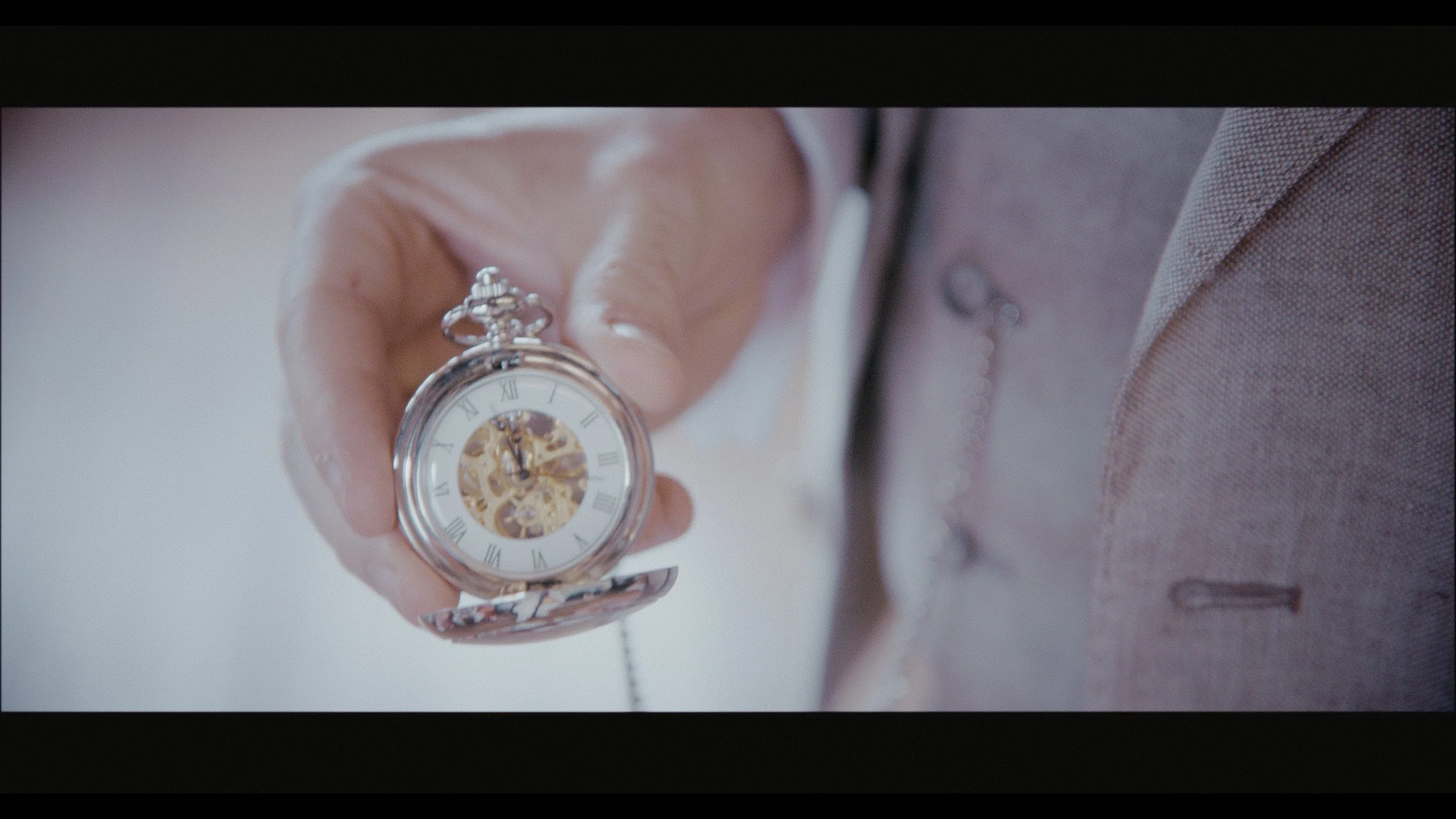 A cinematic close-up captured by Heart Shaped Video, showing a groom holding a vintage silver pocket watch on his wedding day. The elegant timepiece, with visible clockwork and Roman numerals, adds timeless charm and sophistication to the soft, roman