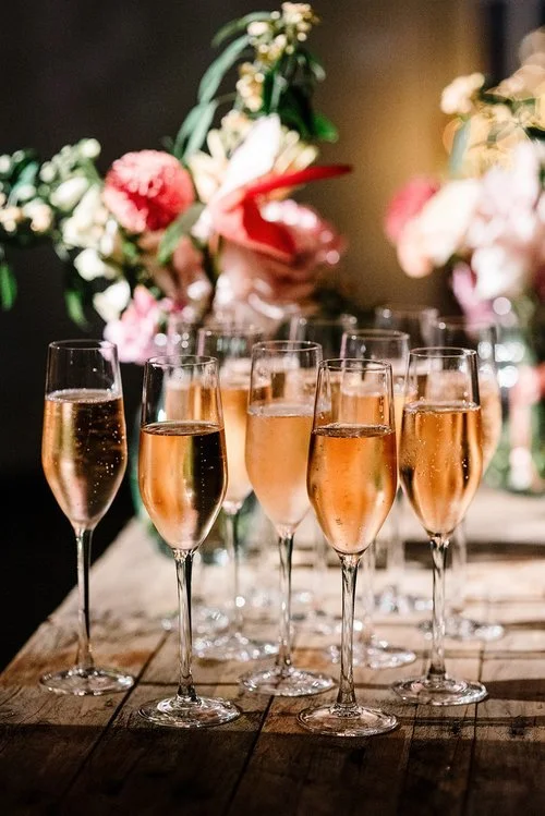 Elegant glasses of sparkling rosé are beautifully arranged on a rustic wooden table, surrounded by soft floral arrangements. This stylish display by Tasting Plates Wedding Caterers showcases their attention to detail and ability to create refined, in