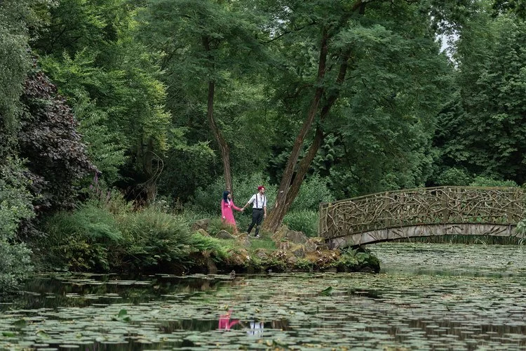 A couple walk hand in hand along a lakeside path surrounded by lush greenery, beautifully photographed by Christy Photography. The ornate wooden bridge, calm water and natural woodland setting create a peaceful, romantic atmosphere that highlights th