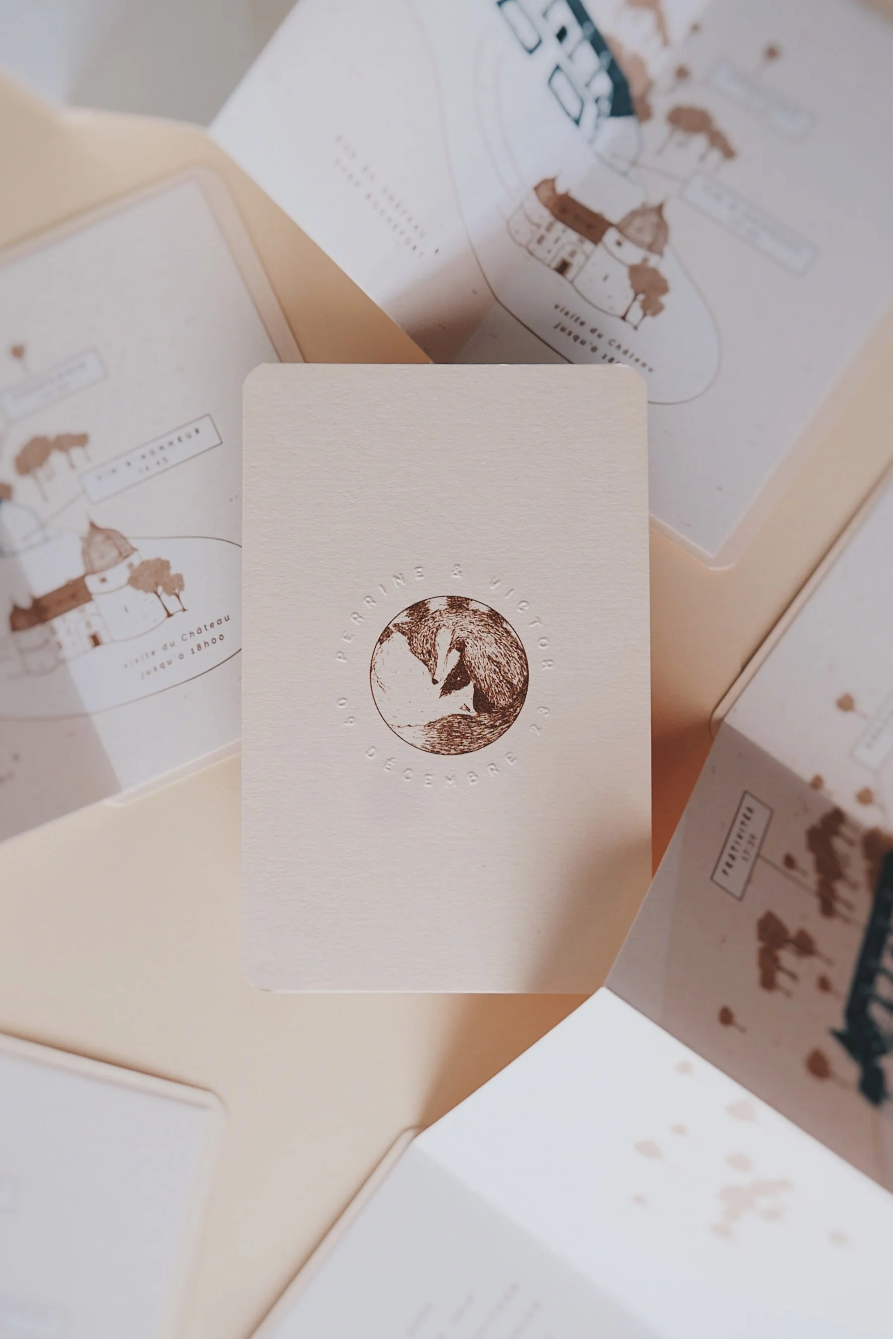 A refined wedding invitation flatlay featuring a soft cream card embossed with a circular illustration and the couple’s names surrounded by elegant custom map inserts in muted pastel tones creating a sophisticated travel inspired aesthetic perfect fo