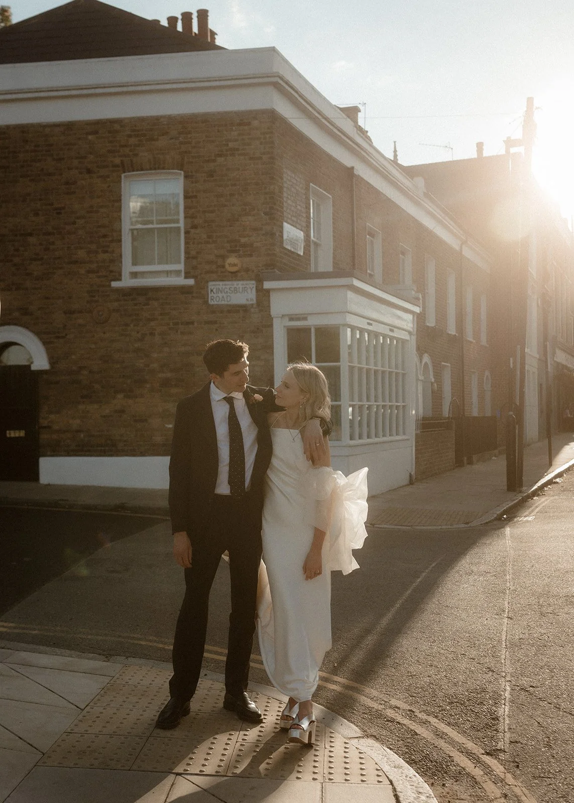 A couple stands on a quiet corner at golden hour, the low sun wrapping the brick terraced houses behind them in warm light as they lean into each other with relaxed affection; the groom in a dark suit rests his arm around the bride’s shoulders while 