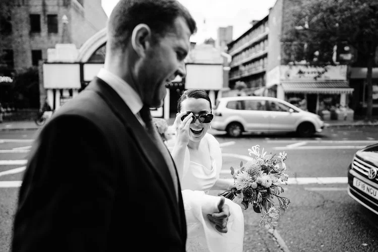A couple share a burst of laughter as they cross a busy London street, the bride playfully lifting her sunglasses while holding a textured bouquet of seasonal flowers. The candid black-and-white moment captures their joy and spontaneity against the b