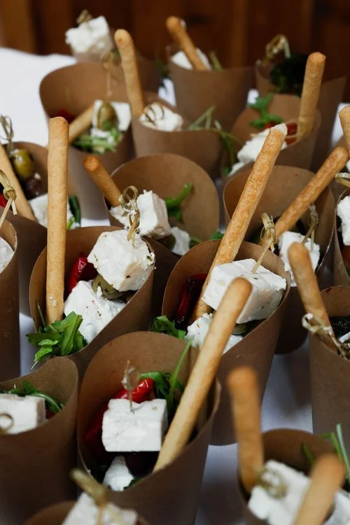 Individual canapé cones filled with feta cheese cubes, roasted peppers, olives, and fresh greens, each topped with a crisp breadstick. This creative appetiser presentation by Regis Banqueting highlights their modern wedding catering style, combining 