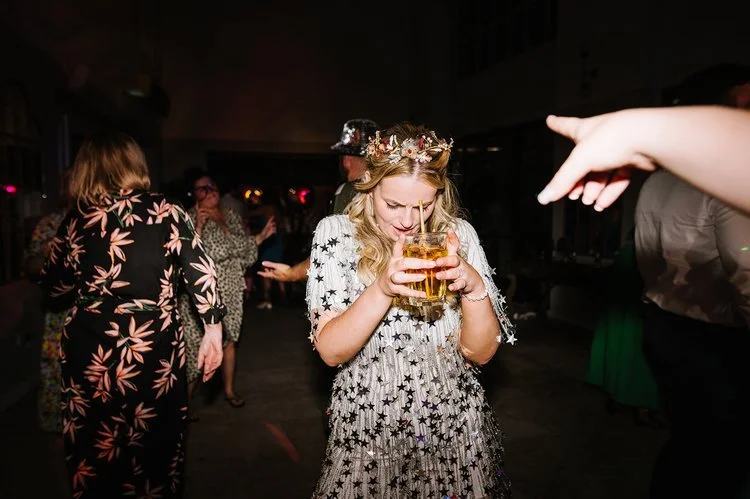 A joyful wedding guest dances and laughs with a drink in hand during a lively wedding reception, surrounded by friends enjoying the party atmosphere. The fun and carefree moment captures the energy and celebration brought by The Best Men wedding DJ.