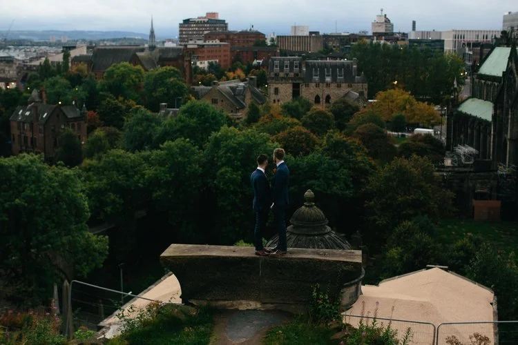 A breathtaking cityscape wedding portrait captured by Mirrorbox Photography, featuring a couple standing together on a historic stone viewpoint overlooking Glasgow’s skyline. Dressed in matching blue suits, they share a quiet moment while surrounded 