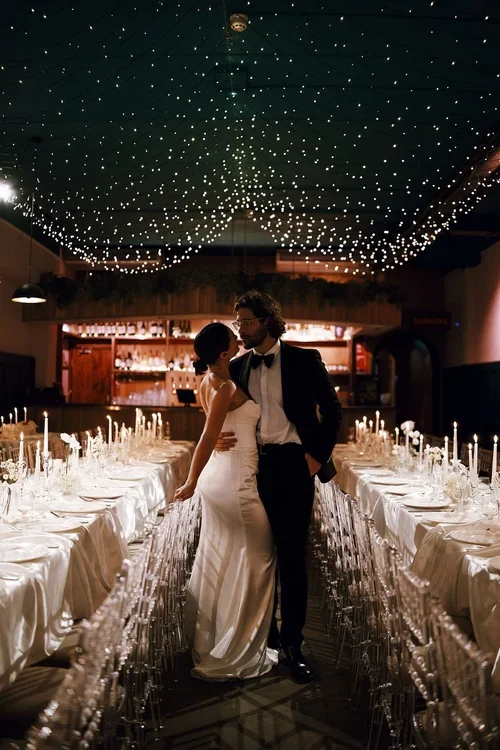 A newlywed couple shares an intimate moment under a canopy of twinkling fairy lights at Colours Hoxton in Shoreditch, London. The romantic wedding setup features long banquet tables draped in satin linens, elegant glassware, and tall taper candles, a