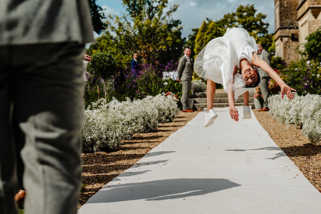 A flower girl launches into an exuberant mid-air flip along the outdoor aisle with her dress billowing around her and arms outstretched while suited guests look on laughing among vibrant garden borders under bright afternoon sun.