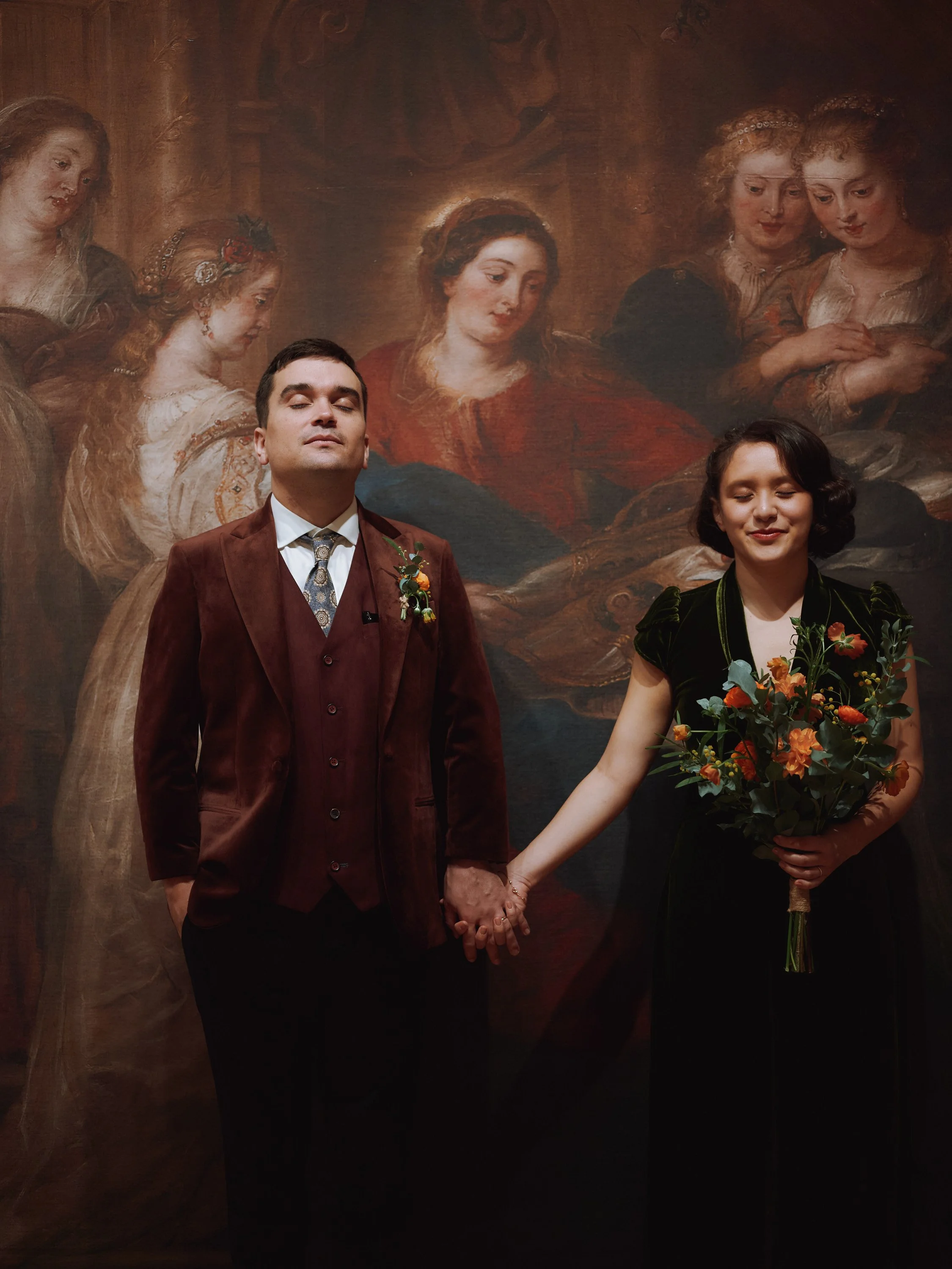 A richly atmospheric wedding portrait captured by Wayne La Photography, showing the couple standing hand in hand before an opulent classical painting. The groom’s deep burgundy suit and composed expression create a striking contrast with the bride’s 