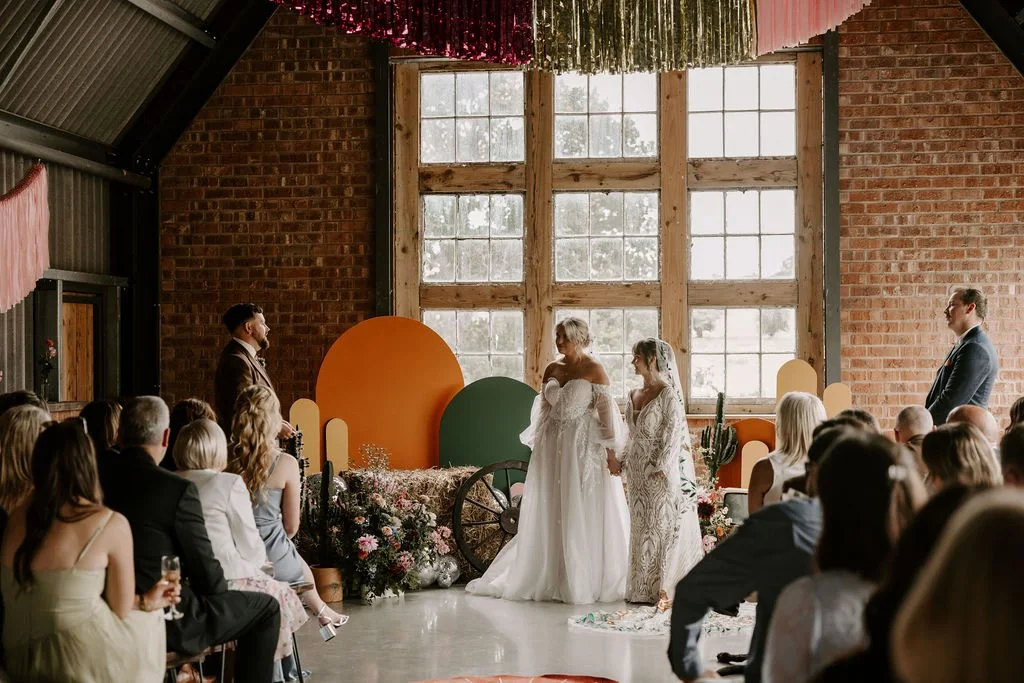 A wedding ceremony at The Giraffe Shed in Wales, featuring two brides standing before a vibrant geometric backdrop of orange, green, and mustard panels beneath a large industrial window. The space blends rustic brick walls with modern, colorful styli