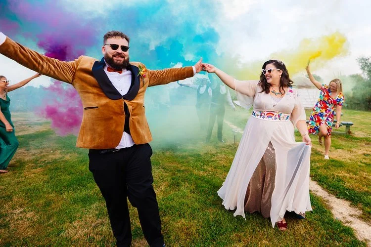 An outdoor wedding celebration featuring a joyous couple dancing through vibrant clouds of coloured smoke. The groom wears a tan velvet tuxedo jacket and sunglasses, while the bride is dressed in a flowing white and gold gown with embroidered detaili