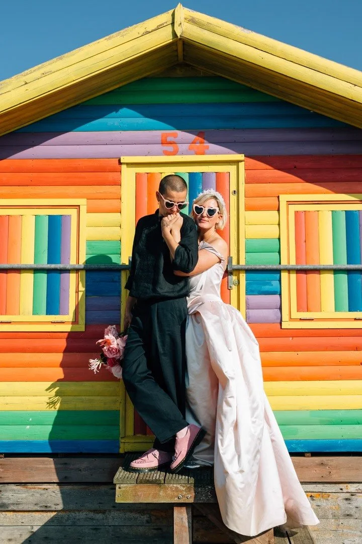 A stylish LGBTQ+ couple pose in front of a brightly painted rainbow beach hut, wearing matching heart-shaped sunglasses. One partner stands confidently in a chic black outfit, holding a bouquet of pink florals, while the other leans against them in a