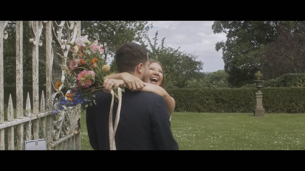 A couple share an unfiltered burst of joy as they embrace beside an ornate weathered gate with the bride’s colourful bouquet spilling vibrant florals across the frame and the surrounding garden adding soft greenery that enhances the carefree romantic