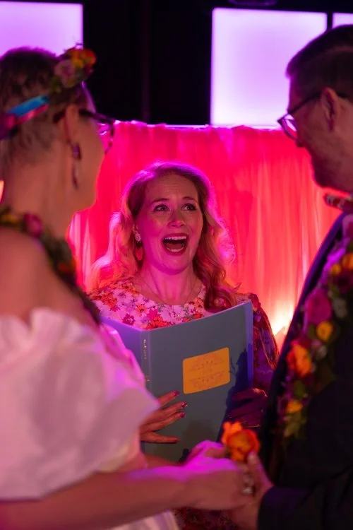 Vibrant wedding ceremony filled with joy and colour, featuring Uno The Joy Celebrant laughing warmly as the couple exchange vows. The scene is illuminated by pink and purple lighting, with the couple wearing floral garlands and bright, creative attir