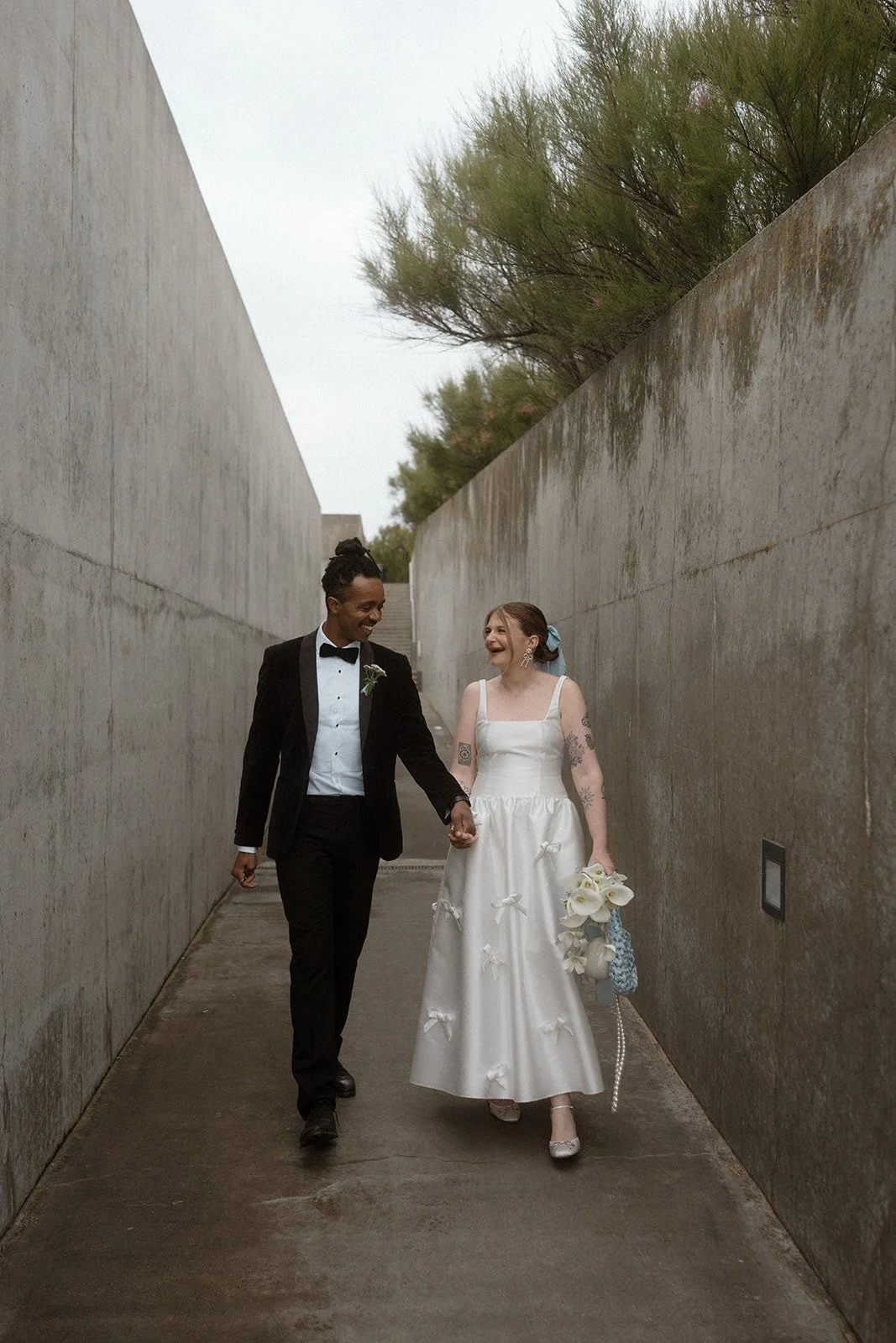 A bride and groom walk hand in hand through a long outdoor concrete corridor with trees visible above the walls, with the groom in a black tuxedo and the bride in a white dress with bow details carrying a bouquet of white flowers.