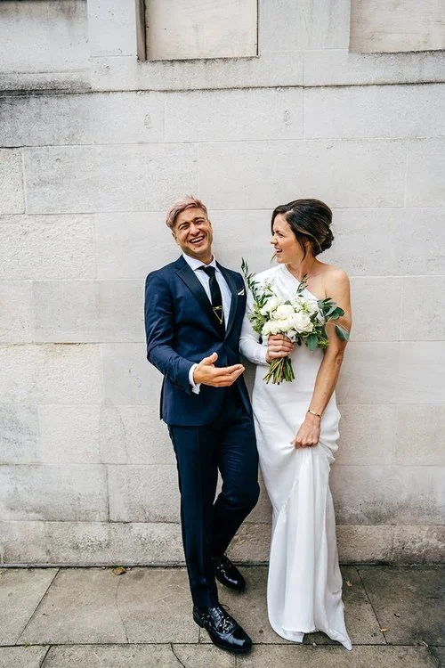A newly married couple share a fun, light-hearted moment as they laugh together against a textured stone wall, beautifully captured by Jordanna Marston Photography. The bride’s elegant white dress and bouquet contrast with the groom’s sharp navy suit