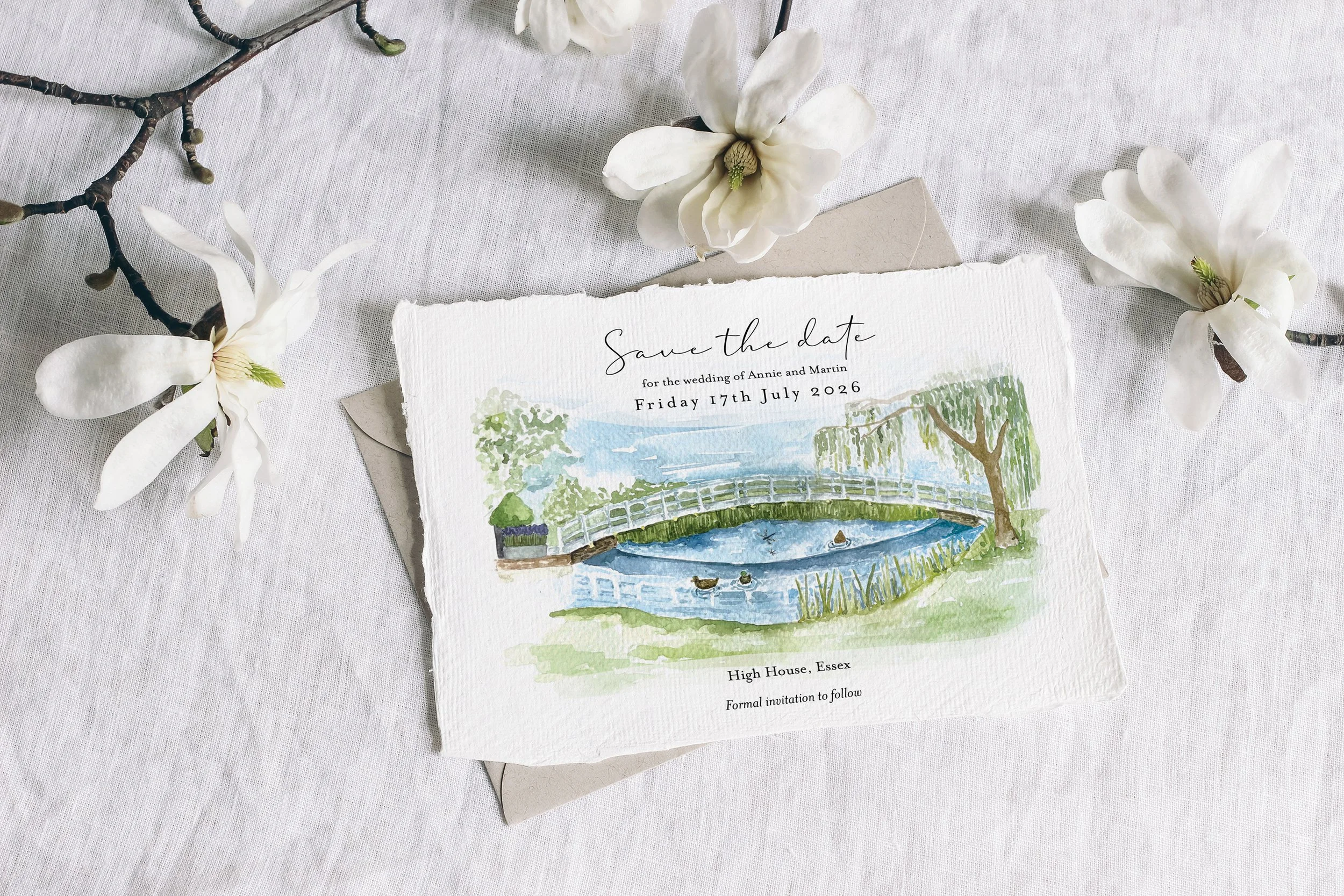 A romantic watercolour save the date card by Feather & Ink Stationery featuring a hand painted scene of a serene pond, wooden bridge and willow trees printed on textured deckle edged paper and styled with magnolia blossoms on soft white linen creatin