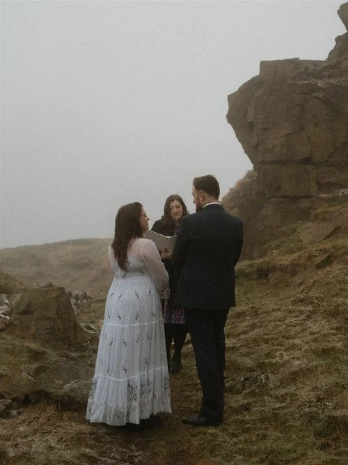 An atmospheric outdoor wedding ceremony taking place on a misty hillside. The couple stands together facing Lauren The Spiritual Celebrant, who is holding a book and smiling warmly as she leads the ceremony. The bride wears a long white dress with de