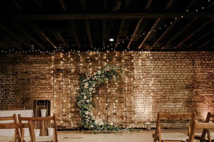 Romantic indoor wedding ceremony featuring a modern crescent floral arch by Catherine Short Florals created with soft roses, lush greenery and natural meadow inspired textures. Set against exposed brick and warm fairy lights, this atmospheric botanic