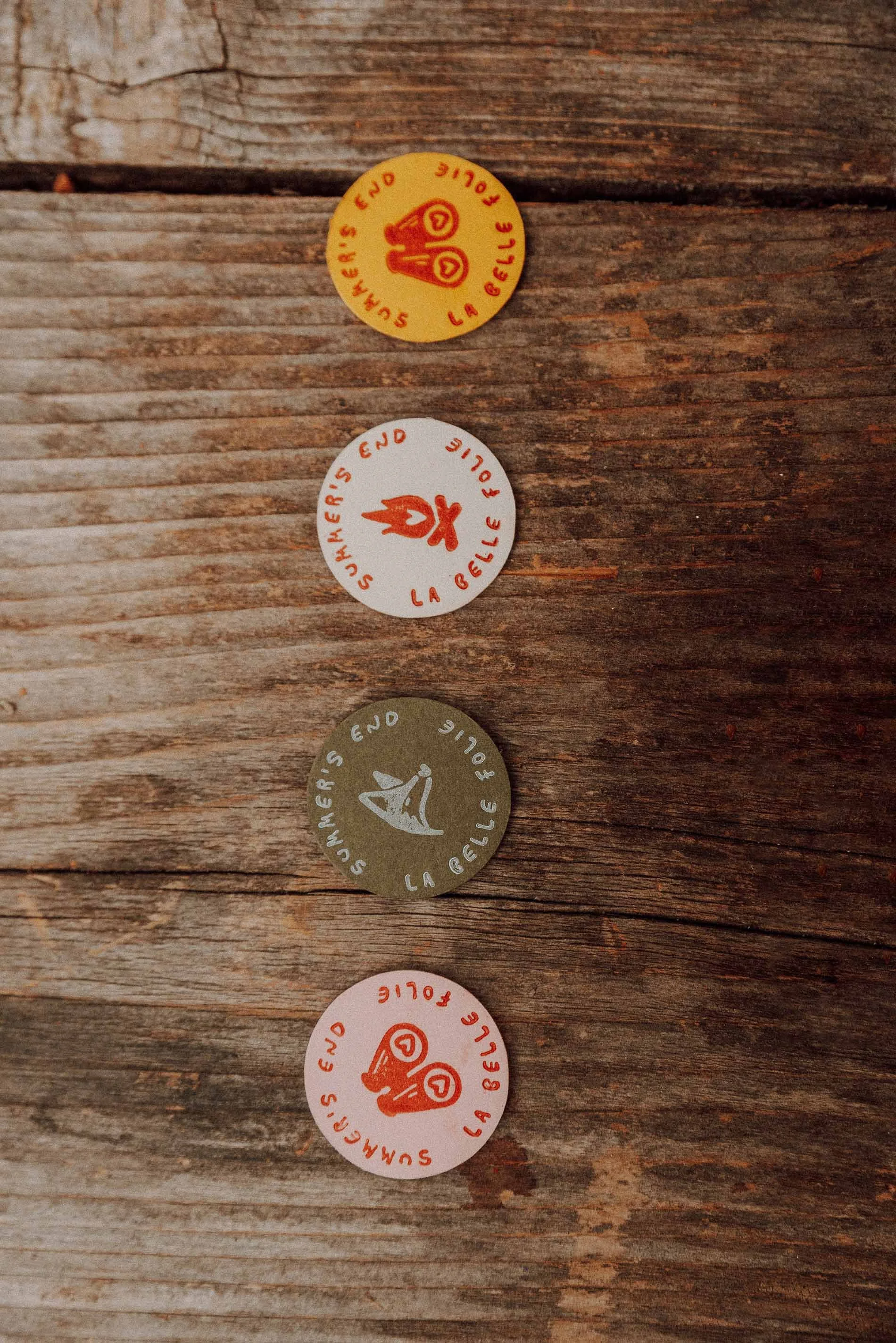 A whimsical wedding detail flatlay featuring four colourful round tokens printed with playful animal illustrations and the wording Summer’s End La Belle Folie arranged vertically on a rustic wooden surface creating a charming Wes Anderson inspired ae