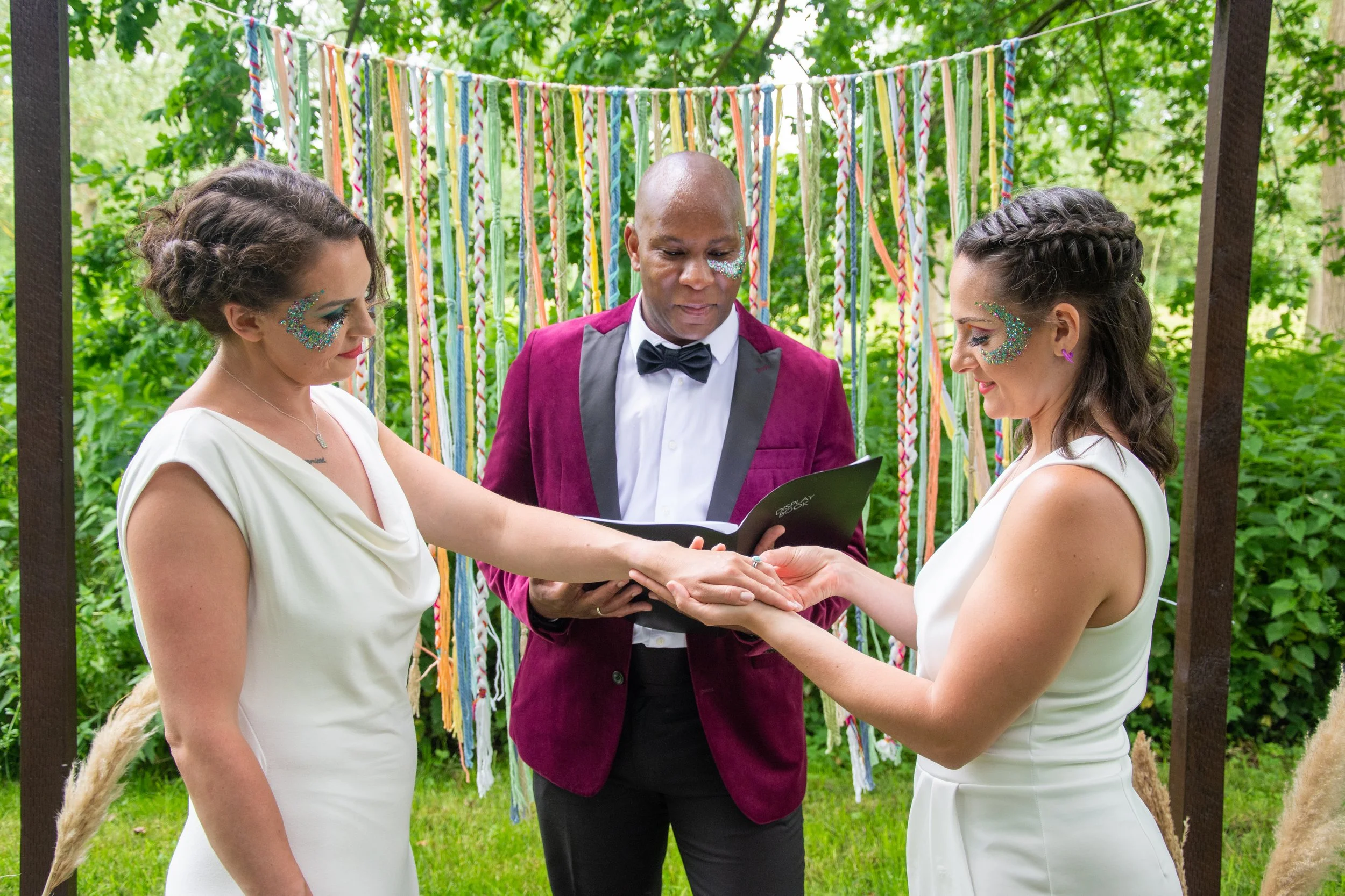Dave Octave Celebrant in a maroon blazer, with 2 brides during a same sex wedding ceremony. One bride is putting a ring on the other ones finger. 