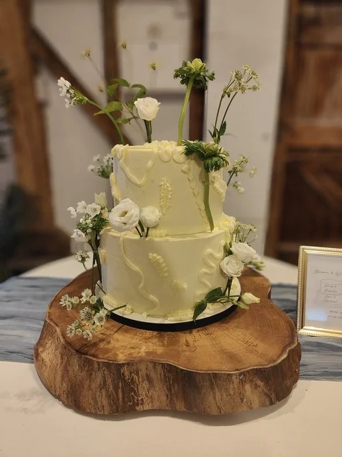 A stunning two-tier buttercream wedding cake by Amber’s Cakes and Treats, featuring a soft pastel yellow finish with delicately piped botanical textures. Fresh white blooms and greenery are artfully arranged to create a natural, organic aesthetic, wh