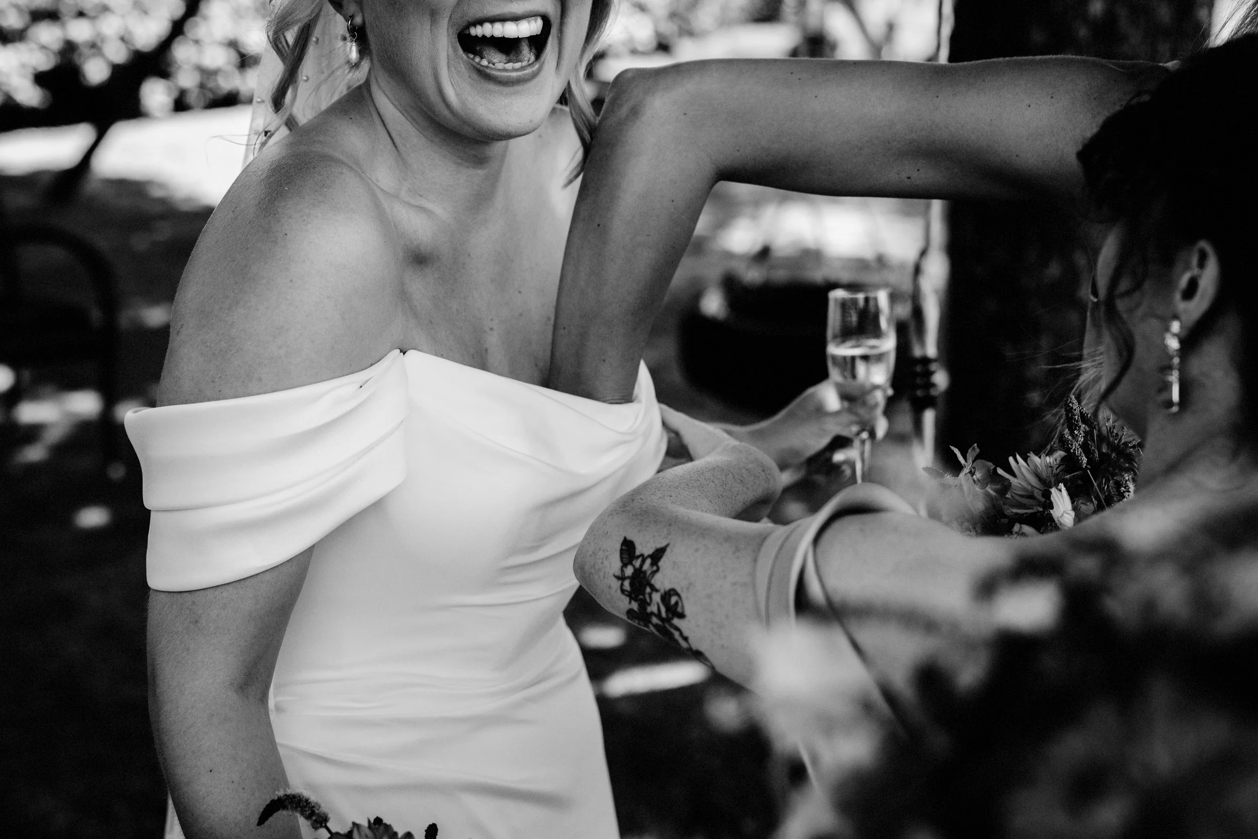 A candid black and white wedding photo captured by Teddy Photo + Film showing two guests sharing a joyful moment during outdoor celebrations. The foreground focuses on a bride in an off-the-shoulder dress laughing as another guest reaches in with a g