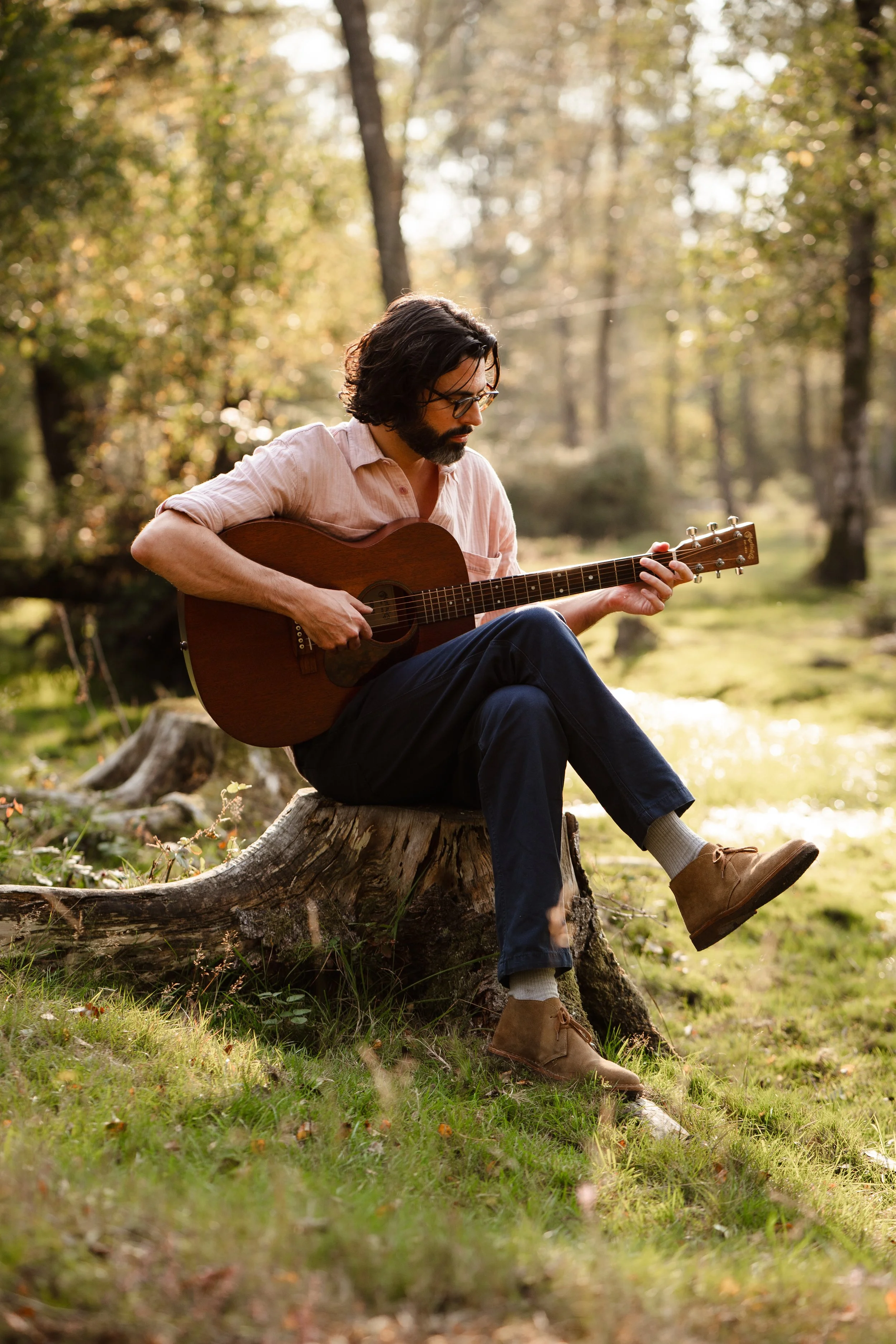 Thomas J Wilman, an acoustic wedding singer and guitarist, performs while seated on a tree stump in the woods, bathed in warm natural light. His relaxed, soulful style makes him perfect for rustic outdoor weddings and intimate acoustic performances. 