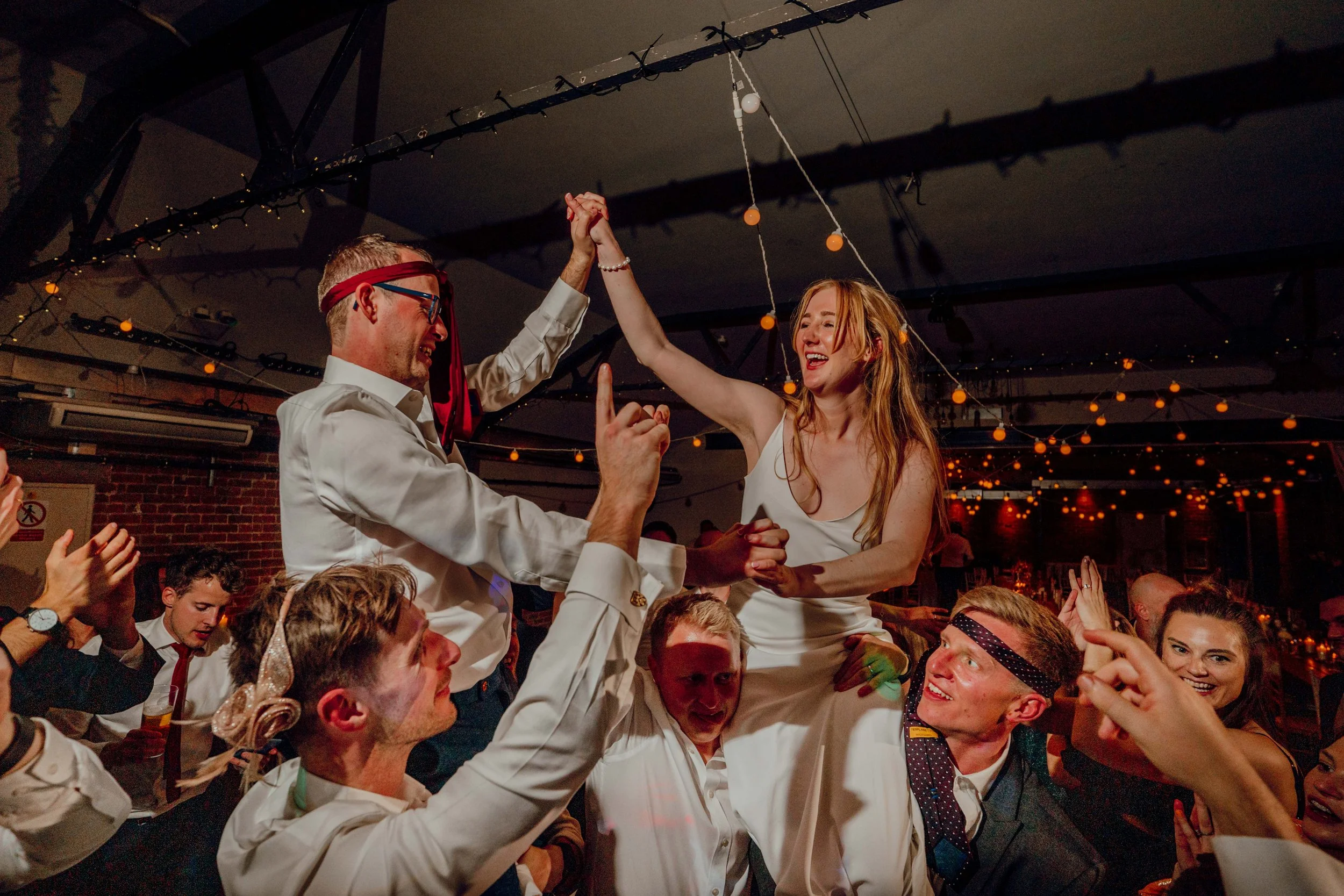 A bride and groom are on the shoulders of their wedding guests at New Craven Hall in Leeds. The photo is dimly lit as it is an evening wedding reception. The dancefloor looks lively.