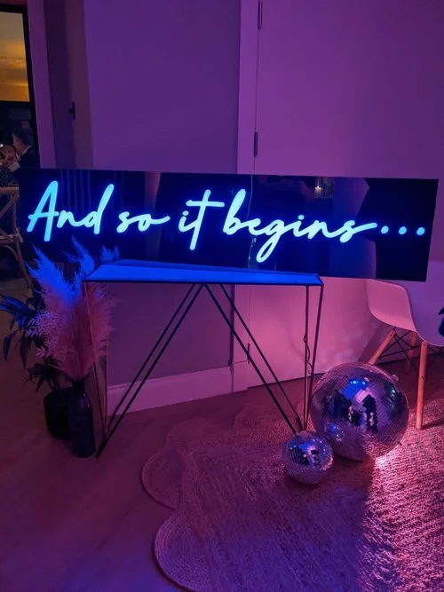 My Colour is Gold blue neon sign reading 'And so it begins…', styled with disco balls and pampas grass for a modern wedding or celebration backdrop.