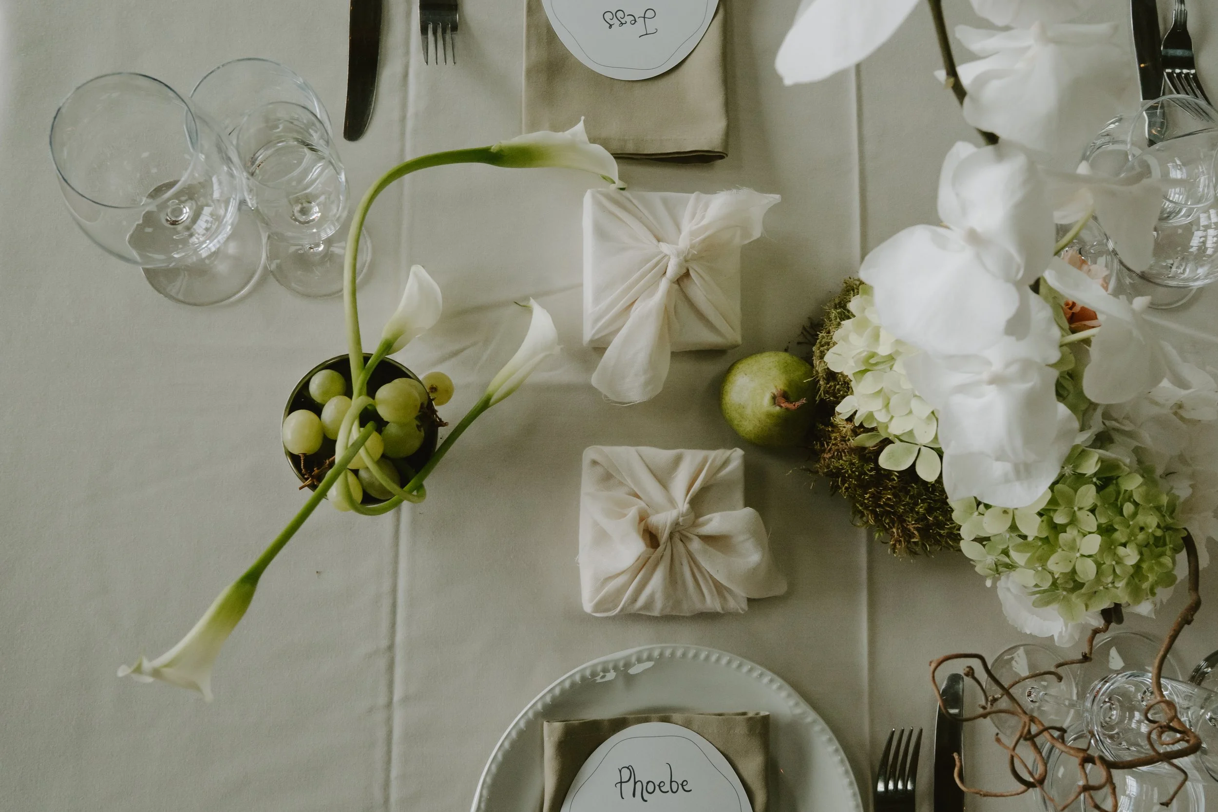 An overhead view of a softly styled wedding table set in muted, natural tones. Pale linen tablecloths are paired with white ceramic plates, clear glassware and neatly folded neutral napkins with handwritten place names. Sculptural floral arrangements
