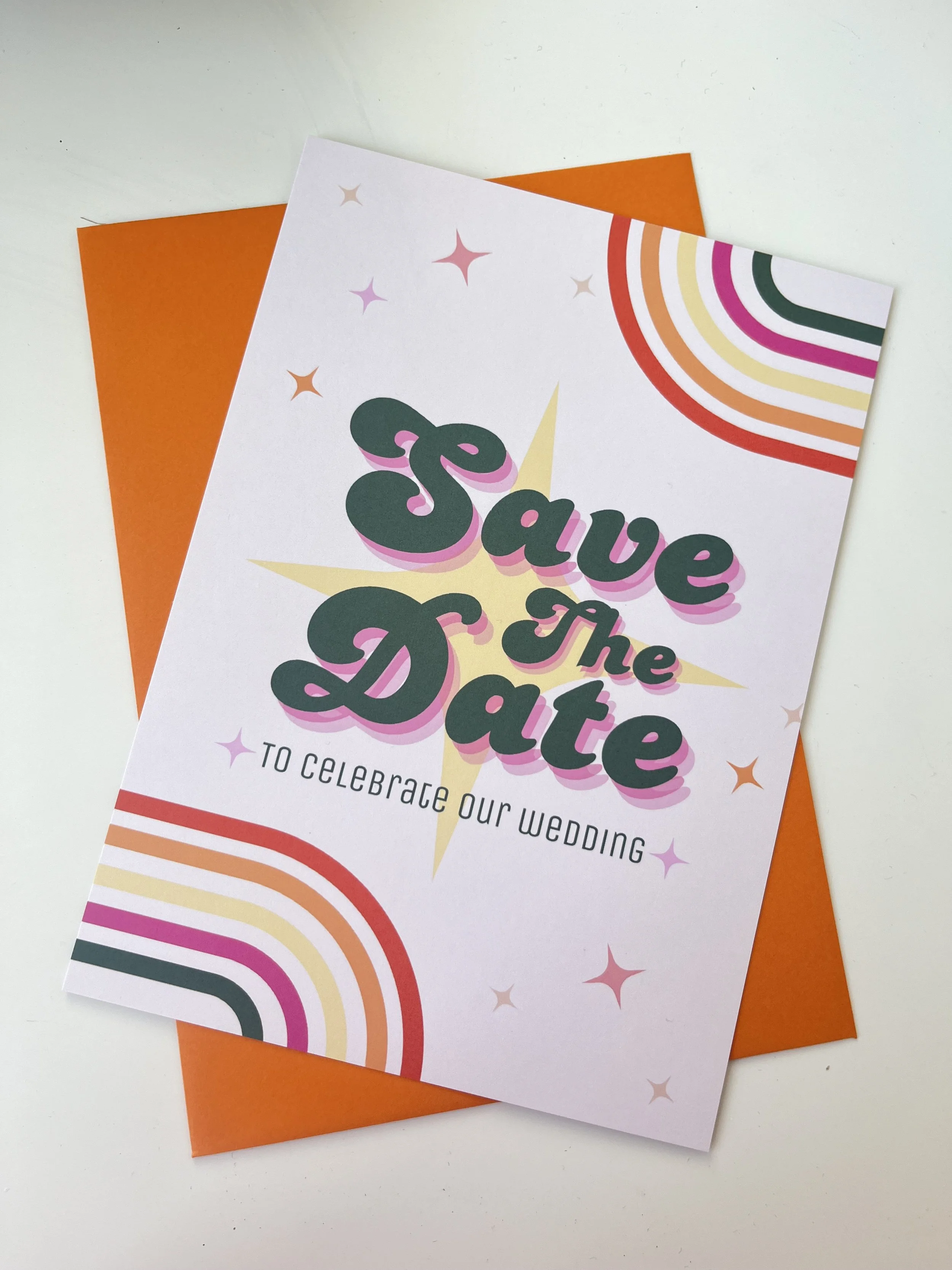 A colourful wedding invitation card is shown layered over a burnt-orange envelope on a light background. The design features bold, retro-style lettering reading “Save the Date” with soft shadowing, set against a pale base with starburst details and c