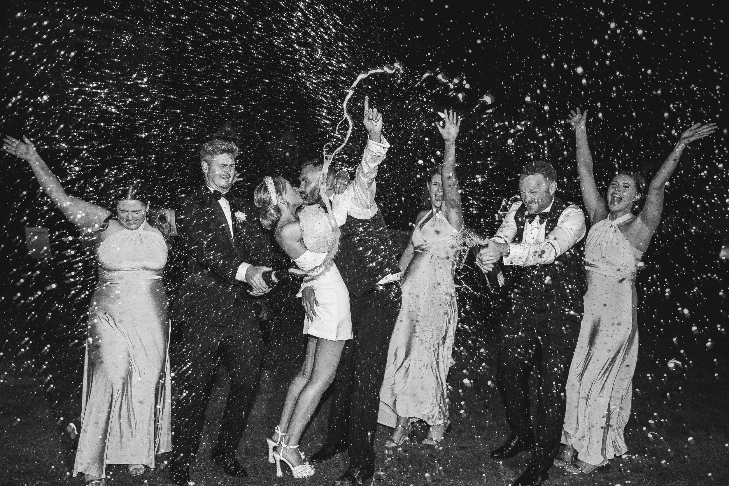 A high-energy black-and-white wedding party moment captured by Gemma Gaskins Photography, featuring a couple kissing as friends surround them, spraying champagne into the night sky. The celebratory scene is full of motion, joy and sparkling party vib