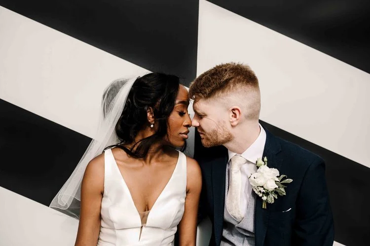 A bold and contemporary wedding portrait captured by Amy Faith Photography, showing the couple leaning gently toward one another against a striking black-and-white geometric wall. The bride’s sleek satin gown and soft veil create a beautiful contrast