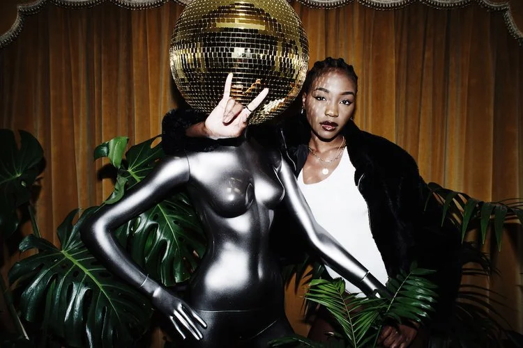 A cool Black woman with her arm around a Disco Queen mannequin from My Colour Is Gold.
