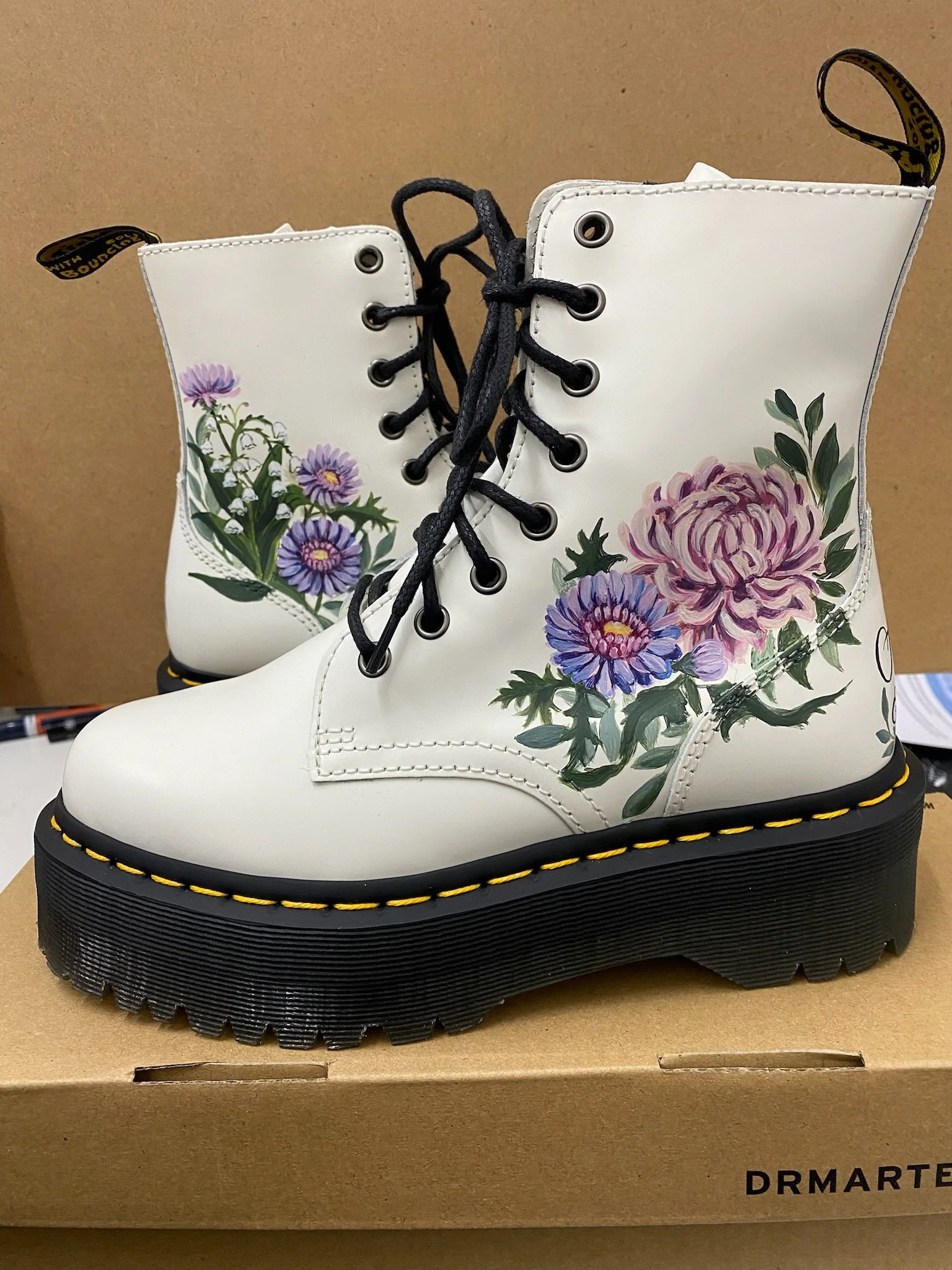 Custom hand-painted leather boots featuring detailed floral illustrations in soft lilac, pink and green tones, set against a clean white base with black laces and chunky soles. The artwork combines fine botanical painting with bold, wearable design, 