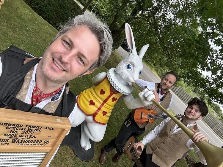 The Ukes of Hazard pose for a playful selfie with a large “White Rabbit” statue from Alice in Wonderland, holding their instruments and grinning for the camera. Dressed in tweed waistcoats, shirts, and red neckerchiefs, the trio exude their signature