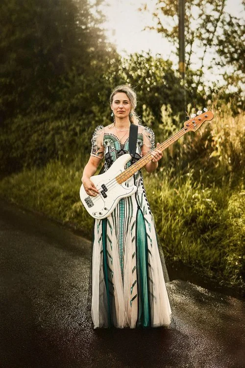 A striking portrait created in Matt Parry Photography’s bold, character-driven documentary style. A woman stands confidently on a wet country road, holding a white electric bass guitar as if ready to perform. She wears an ornate, intricately patterne