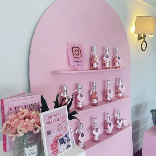 A chic pink display by The Pink Drinks Van featuring mini bottles of rosé prosecco arranged on pastel pink shelves. Styled with floral accents, branded signage, and a Prosecco cocktail recipe book, this elegant setup perfectly captures The Pink Drink