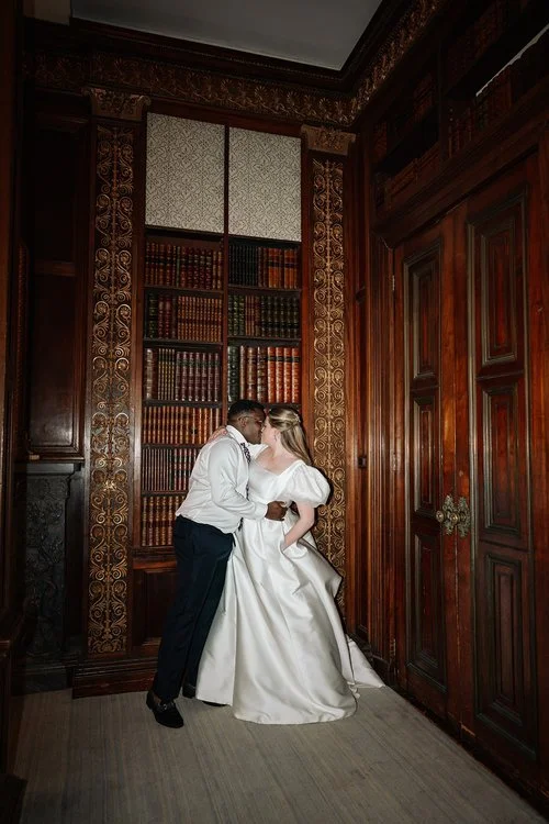 A couple share a quiet kiss in an ornate wood panelled library, framed by towering bookshelves filled with rich tones and intricate detailing. The bride’s dramatic satin gown with voluminous sleeves contrasts beautifully with the warm, historic setti