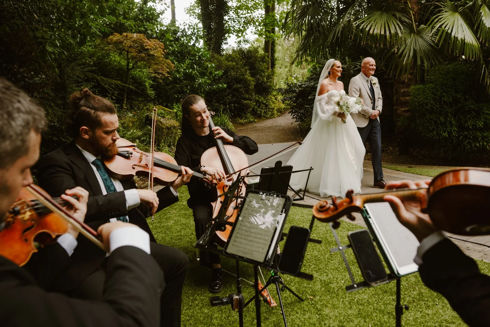 A candid wedding ceremony moment captured by Bluebell Photo Studio, featuring a bride walking down an outdoor garden aisle with her escort while a live string quartet performs. Surrounded by lush greenery and natural light, this elegant wedding photo